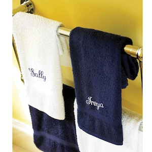 以下が含まれることがあります： クロムタオルラックに掛けられた2つのパーソナライズされたバスタオル。1つのタオルは白で、名前「Sally」がネイビーブルーで刺繍されています。もう1つのタオルはネイビーブルーで、名前「Freya」が白で刺繍されています。