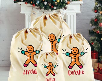 Könnte beinhalten: Ein Set aus vier beigefarbenen Geschenkbeuteln mit Kordelzug und Lebkuchenmann-Designs. Die Lebkuchenmänner haben orangefarbene Körper, rote und blaue Akzente und sind mit Namen in Rot beschriftet. Ein Weihnachtsbaum und Girlanden sind im Hintergrund.
