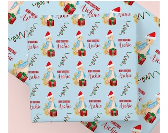 May include: Light blue wrapping paper with a repeating pattern of a bunny wearing a Santa hat, holding a gift, and the text "Merry Christmas Archie". Green and red ribbons and floral accents complete the design.