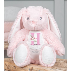 Puede incluir: Un peluche rosa con forma de conejo, con orejas largas y detalles blancos. El conejo tiene un diseño personalizado en el pecho con el nombre "Harper" y la pregunta "¿Quieres ser mi niña de las flores?"