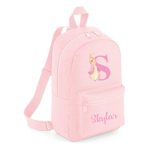 Könnte beinhalten: Rosa Kinderrucksack mit Peter Rabbit-Motiv und dem Buchstaben "S" in Rosa. Der Rucksack hat ein Reißverschlussfach und der Name "Skylar" ist auf der Vorderseite aufgedruckt.