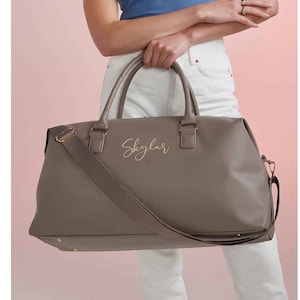 Könnte beinhalten: Eine graue Ledertasche mit goldfarbenem Reißverschluss und goldfarbenem Trageriemen. Die Tasche trägt den Namen "Skylar" in goldenen Lettern.