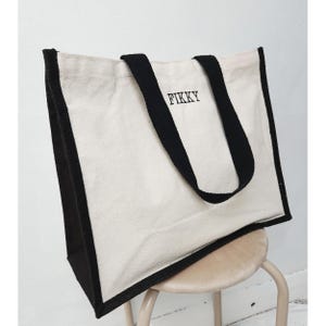 Puede incluir: Un bolso tote de lona color crema con ribetes y asas negras. La palabra "FIKKY" está impresa en negro en la parte delantera. El bolso está sobre un taburete pequeño y claro. Diseño sencillo y funcional.
