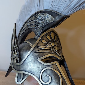 Op de afbeelding: Een metallic donkergrijze en goudkleurige helm met een opvallende witte kam. De helm is voorzien van gedetailleerde reliëfmotieven van vleugels aan de zijkanten en een gestileerd bloemmotief op het voorhoofd, met een puntige neusbeschermer.