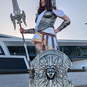 May include: A person in a detailed cosplay of a Greek warrior, complete with a spear, shield, and ornate armor. The shield features a Medusa design. The costume is predominantly silver, white, and brown.