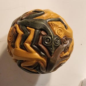 Può includere: Una scultura sferica in legno con una superficie testurizzata. La scultura è dipinta in tonalità di marrone, verde e giallo. La scultura rappresenta più figure intrecciate tra loro.