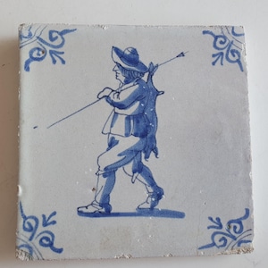 Könnte beinhalten: Eine blaue und weiße Delft-Kachel mit der Darstellung eines Mannes mit Hut, der einen langen Stock trägt. Die Kachel hat einen dekorativen Rand.