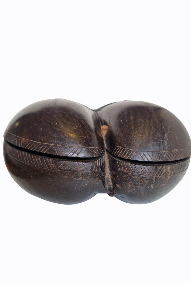 Op de afbeelding: Donkerbruine, dubbelgelobde houten doos met een gesneden geometrisch ontwerp rond de rand van elke lob. De doos lijkt met de hand gesneden te zijn en heeft een natuurlijke, rustieke uitstraling. De twee helften zijn in het midden verbonden.