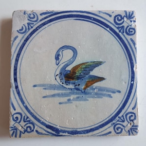 Puede incluir: Un azulejo de cerámica blanco con un borde azul y blanco y una imagen pintada a mano de un cisne azul con detalles marrones y verdes en el centro.