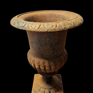 May include: A weathered, cast iron urn with a wide, decorative rim and a bulbous midsection. The urn sits on a square base and has a dark, rusty brown color, suggesting outdoor use. The design features ornate detailing.