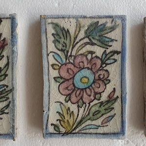 May include: Three rectangular ceramic tiles with floral designs. Each tile features a different flower arrangement in shades of pink, yellow, blue, and green, set against a cream-colored background. The tiles are framed with a blue border.