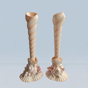Puede incluir: Dos candelabros con forma de concha. Los candelabros tienen un diseño en espiral y son de color beige claro. La base de cada soporte está decorada con conchas marinas y coral en tonos rosas y beige.