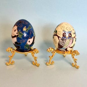 Puede incluir: Dos huevos de porcelana decorativos en soportes dorados. El huevo de la izquierda presenta un fondo azul con dos pandas y hojas verdes. El huevo de la derecha presenta un fondo blanco con un búho con plumas azules, verdes y rosas.