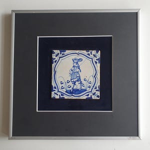 May include: A framed blue and white Delft tile depicting a man in a feathered hat and a patterned coat standing in a garden setting.