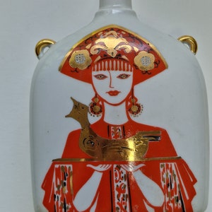 Op de afbeelding: Een witte keramische fles met een rood en gouden ontwerp. De fles toont een vrouw in een rode jurk en een gouden hoofdtooi die een gouden vogel vasthoudt.