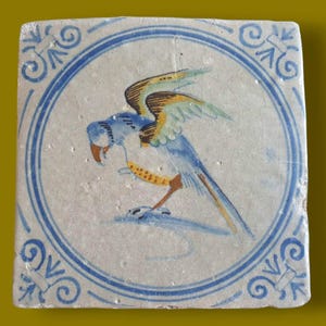 Puede incluir: Una baldosa de cerámica blanca con un pájaro azul y amarillo con las alas extendidas. La baldosa tiene un borde azul y blanco con un patrón decorativo.