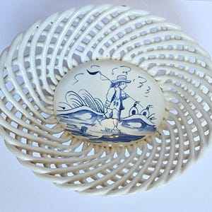 May include: White ceramic basket with a blue and white hand-painted design of a person with a flag and two birds. The basket is woven in a spiral pattern.