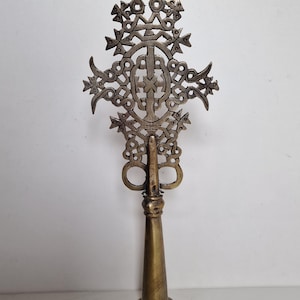 Può includere: Un'antica croce in metallo ornata con un intricato design traforato. La croce presenta un motivo dettagliato di cerchi, elementi floreali e un motivo a croce centrale. La base è a forma di cono color ottone.