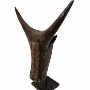 Puede incluir: Escultura de cabeza de toro de madera tallada de color marrón oscuro con cuernos largos curvados hacia arriba. La escultura presenta detalles decorativos rojos y dorados en el hocico y está montada sobre un soporte de metal negro. Los cuernos tienen detalles plateados.