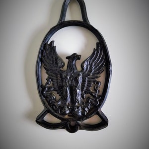 Könnte beinhalten: Schwarzer Wandhaken aus Gusseisen mit Adlermotiv. Der Adler ist in erhabener Form dargestellt und hat ausgebreitete Flügel. Der Haken hat oben eine Schlaufe zum Aufhängen.