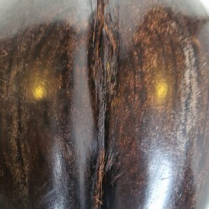 Op de afbeelding: Close-up van een donkerbruin, gepolijst houten object met een verticale, getextureerde naad. De houtnerf is zichtbaar en het oppervlak reflecteert licht, waardoor highlights ontstaan. Het object heeft een gladde, gebogen vorm.