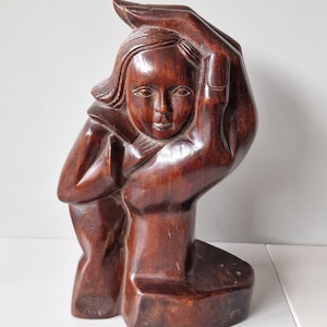 Puede incluir: Una escultura de madera marrón oscuro de una persona con las manos juntas sobre la cabeza. La escultura está tallada en un estilo estilizado y abstracto.