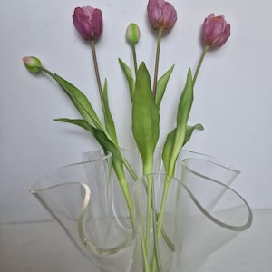 以下が含まれることがあります： 波打つようなフリルデザインの透明なプラスチックの花瓶に、緑の葉のついたピンクのチューリップのブーケが活けられています。