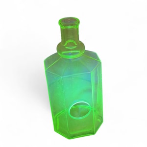 Op de afbeelding: Een neon groene glazen fles met een geometrisch ontwerp. De fles heeft een zeshoekige body en een afgeronde hals. Een ovale opening is zichtbaar aan de zijkant. De fles is ongeveer 15 cm hoog.