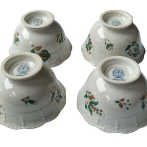 May include: Four white porcelain bowls with a scalloped edge and floral design in green, brown, and gold. Each bowl has a circular base with a blue and white emblem. The bowls are arranged with their bases facing up.