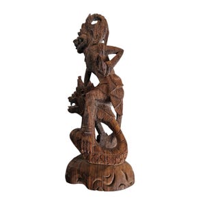 May include: A dark brown wooden sculpture of a mythical creature with a human-like figure riding on its back. The creature has a long tail and a dragon-like head. The sculpture is carved with intricate details and is a traditional Balinese art piece.