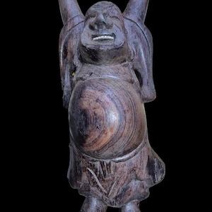 May include: A wooden statue of a laughing Buddha with arms raised. The statue is carved from dark brown wood, with visible grain patterns. The Buddha has a large belly and a smiling face, with a detailed carving of the mouth and teeth. The statue is standing on a black background.