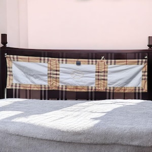 Puede incluir: Una cabecera de madera marrón con un panel de tela a cuadros beige y marrón con tres bolsillos de tela blanca. El panel de tela tiene un pequeño logotipo bordado en el bolsillo central.