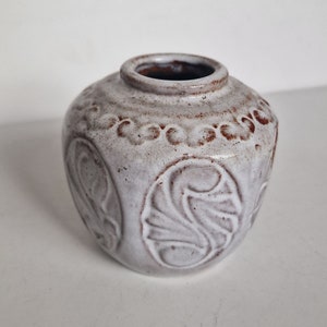 Puede incluir: Un pequeño jarrón redondo de cerámica blanquecina con superficie texturizada y diseños florales en relieve. El jarrón tiene interior y borde marrón oscuro, adecuado para flores pequeñas o como objeto decorativo.