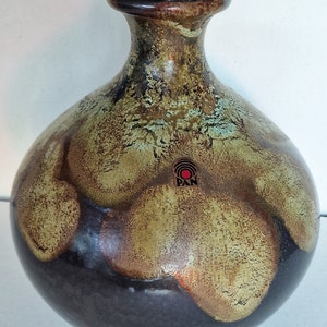 May include: A brown and green ceramic vase with a wide base and a narrow neck. The vase has a mottled glaze with a black base and a green and brown top. The vase has a small, round sticker on the side that says "PAN".