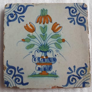 Könnte beinhalten: Eine antike holländische Delft-Kachel mit einem blauen und weißen Blumendesign. Die Kachel zeigt eine Vase mit orangefarbenen und grünen Blumen.