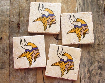 May include: Set of four square stone coasters. Each coaster features a Viking head logo in yellow, purple, and black. The coasters are beige and rest on a wooden surface.
