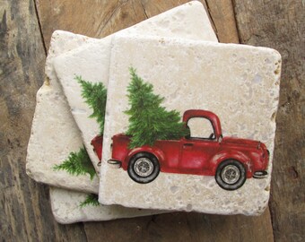 May include: A set of three square stone coasters. Each coaster displays a red vintage truck transporting a green Christmas tree. The coasters have a natural, beige stone appearance, ideal for festive home decor.