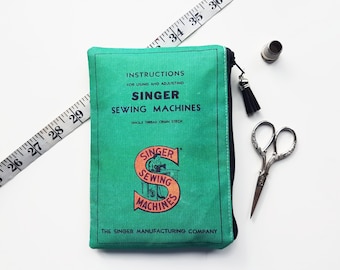 May include: A green zippered pouch with a vintage Singer Sewing Machines logo and instructions for using and adjusting the machines. The pouch is on a white surface with a measuring tape, a thimble, and a pair of small scissors.