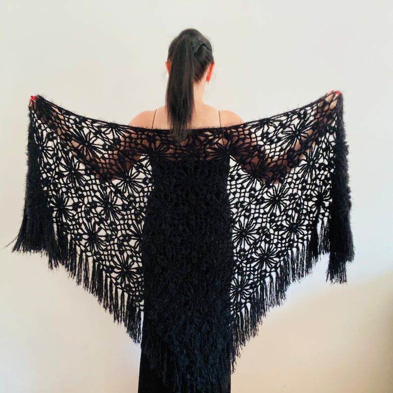 May include: A black shawl with a floral lace pattern and fringe. The shawl is held open, displaying the intricate design and texture. The garment appears to be made of a soft, possibly knitted or crocheted material.
