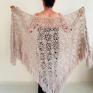May include: A beige crocheted shawl with a floral pattern and fringe trim. The shawl is triangular in shape and is being held up to display the intricate openwork design. The color is a soft, neutral tone.