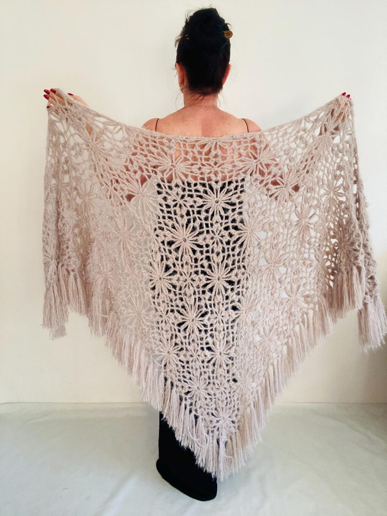 May include: A beige crocheted shawl with a floral pattern and fringe trim. The shawl is triangular in shape and is being held up to display the intricate openwork design. The color is a soft, neutral tone.