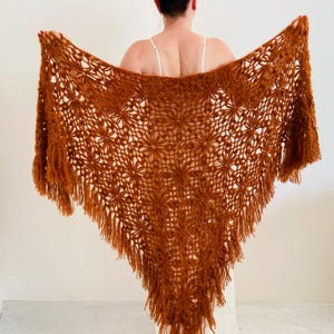 May include: A rust-colored, open-weave shawl with a floral pattern and fringe. The shawl is held open, showcasing its triangular shape and intricate design. The warm, earthy tones suggest a handmade or vintage aesthetic.