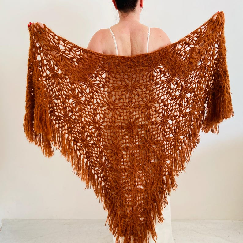 May include: A rust-colored, open-weave shawl with a floral pattern and fringe. The shawl is held open, showcasing its triangular shape and intricate design. The warm, earthy tones suggest a handmade or vintage aesthetic.
