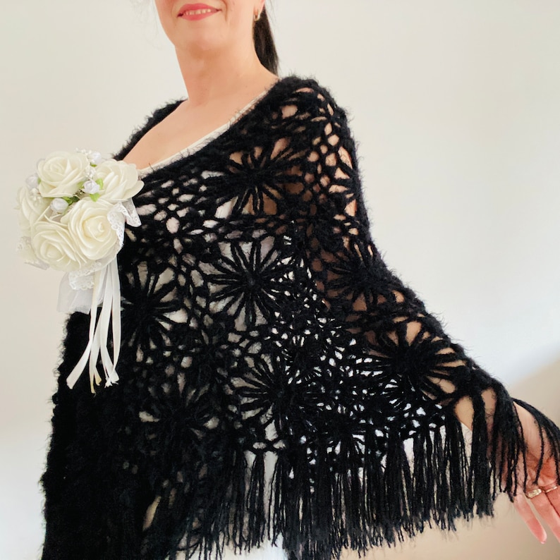 May include: A black, open-weave crochet shawl with a floral pattern and fringe. The shawl is draped over the shoulders and is paired with a white bouquet of roses.
