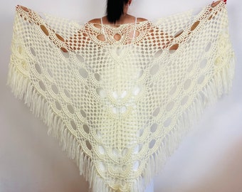 May include: A cream-colored triangular crochet shawl featuring an intricate openwork pattern of repeating circular and fan-shaped motifs. The edges of the shawl are finished with long, straight fringe tassels that hang along the bottom border.