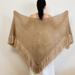 May include: A tan-colored knitted shawl with fringe along the edges. The shawl is triangular in shape and is being held up to display its full form. The fringe is a lighter shade of tan than the main body of the shawl.