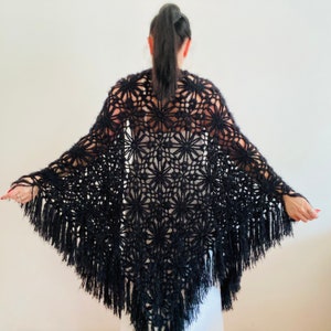 May include: A black, open-weave shawl with a floral pattern and fringe. The shawl is made of a soft, textured yarn and is draped over the shoulders. The fringe hangs down along the bottom edge.