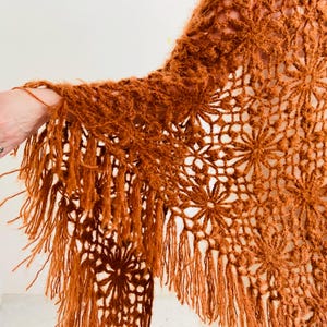 May include: A rust-colored, open-weave shawl with a floral pattern and fringe. The shawl is made of a soft, textured yarn. The fringe hangs from the bottom edge of the shawl. The shawl is draped over a person's arm.