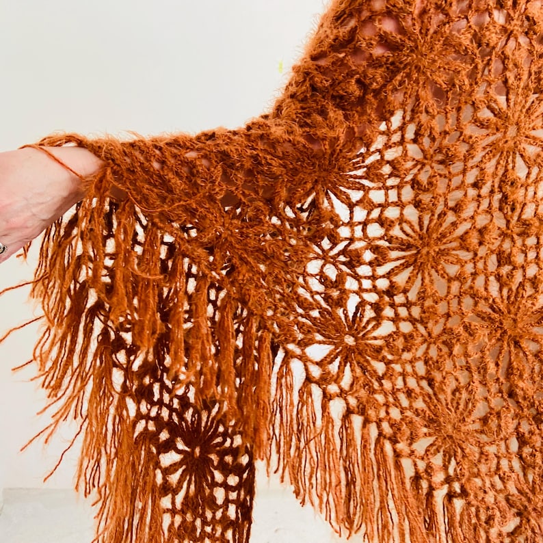 May include: A rust-colored, open-weave shawl with a floral pattern and fringe. The shawl is made of a soft, textured yarn. The fringe hangs from the bottom edge of the shawl. The shawl is draped over a person's arm.