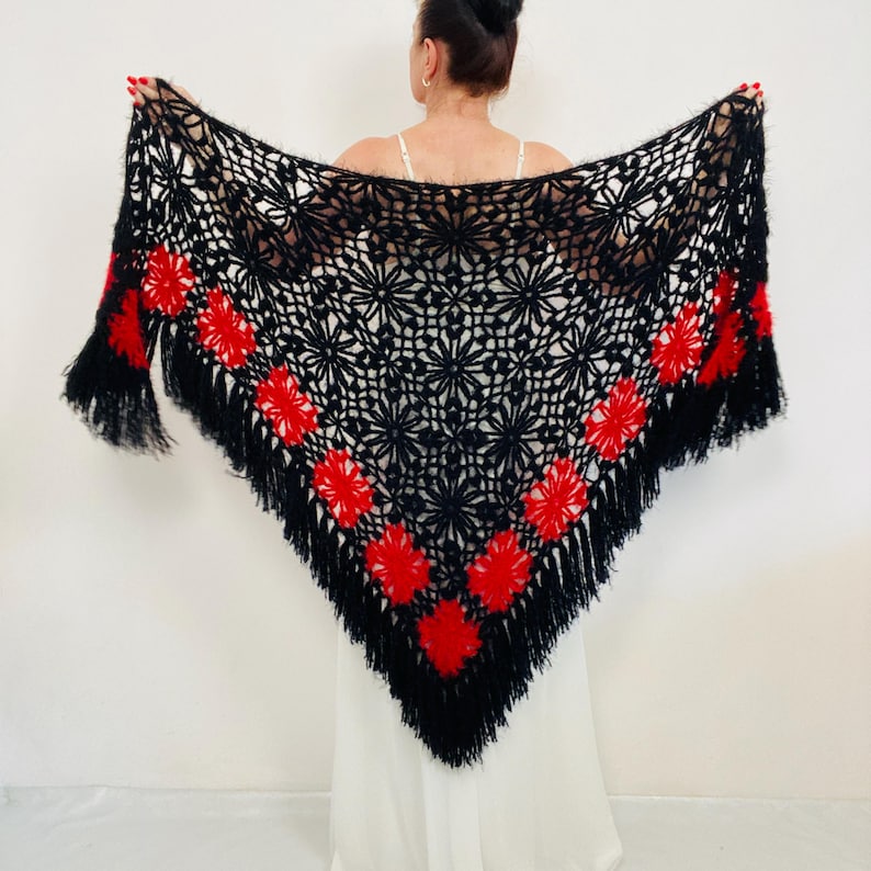 May include: A black crochet shawl with red floral accents and fringe. The shawl is held up, showcasing its intricate openwork design. The red flowers are evenly spaced along the edges, adding a pop of color to the dark background. The shawl is a triangular shape.